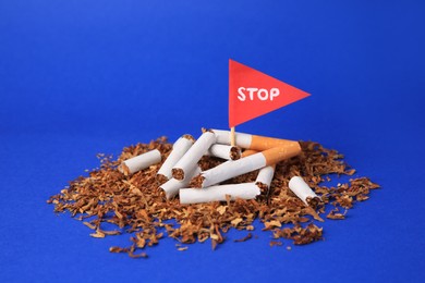 Photo of Broken cigarettes, red flag with word Stop and tobacco on blue background, closeup. Quitting smoking concept