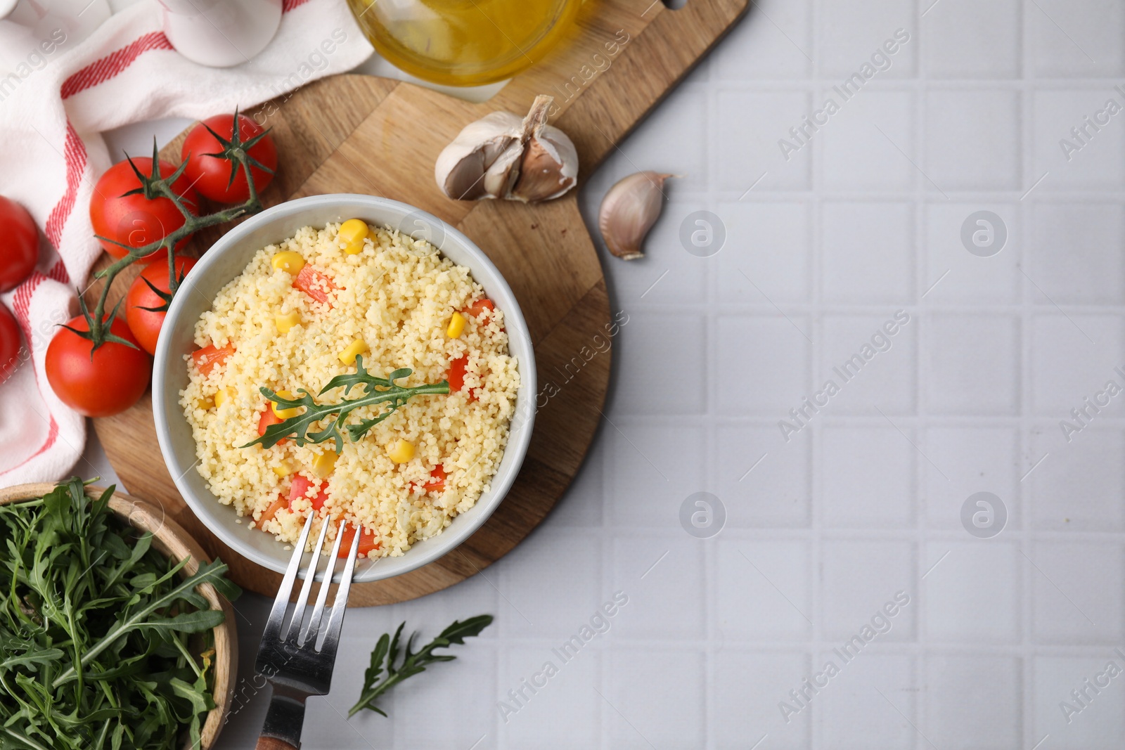 Photo of Tasty couscous with pepper, corn and arugula served on white tiled table, flat lay. Space for text