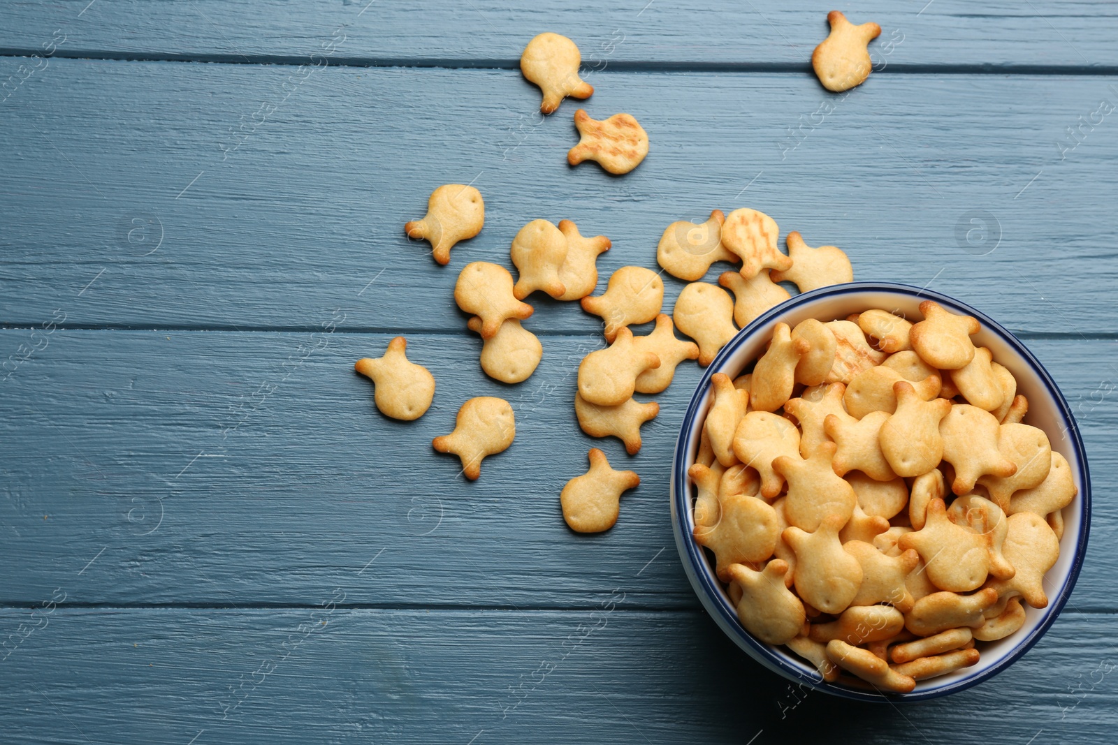 Photo of Delicious goldfish crackers in bowl on blue wooden table, flat lay