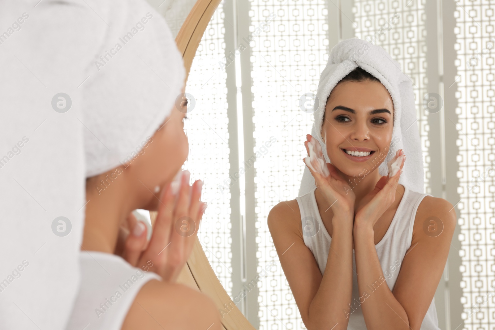 Photo of Happy young woman applying cleansing foam onto face near mirror in bathroom