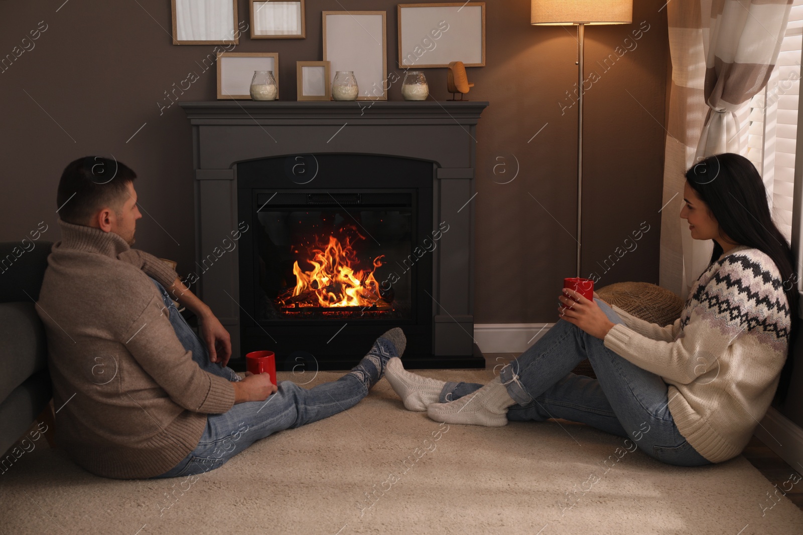 Photo of Lovely couple with cups of hot drink spending time together near fireplace at home