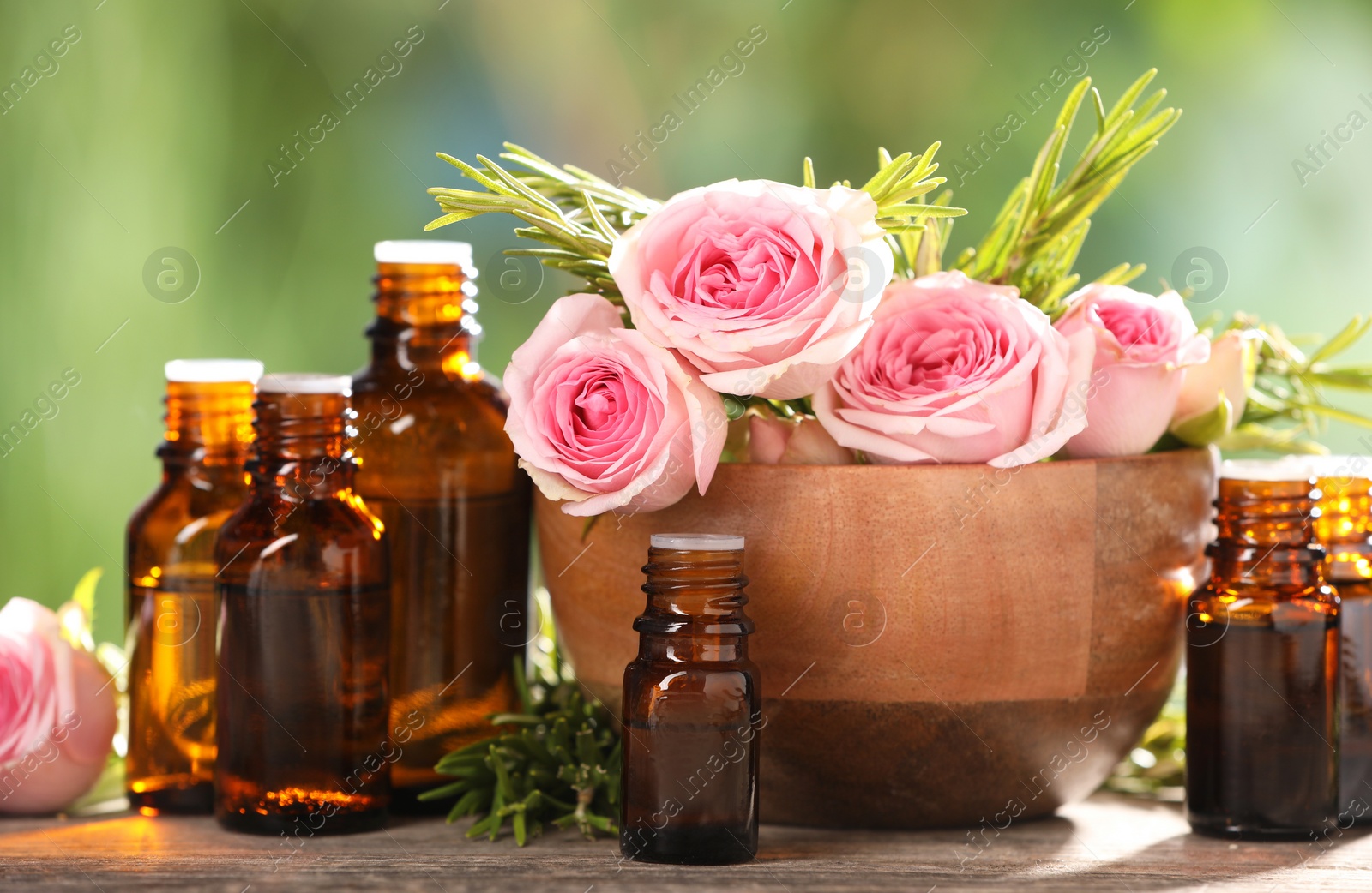 Photo of Bottles with essential oils, roses and rosemary on wooden table against blurred green background, closeup