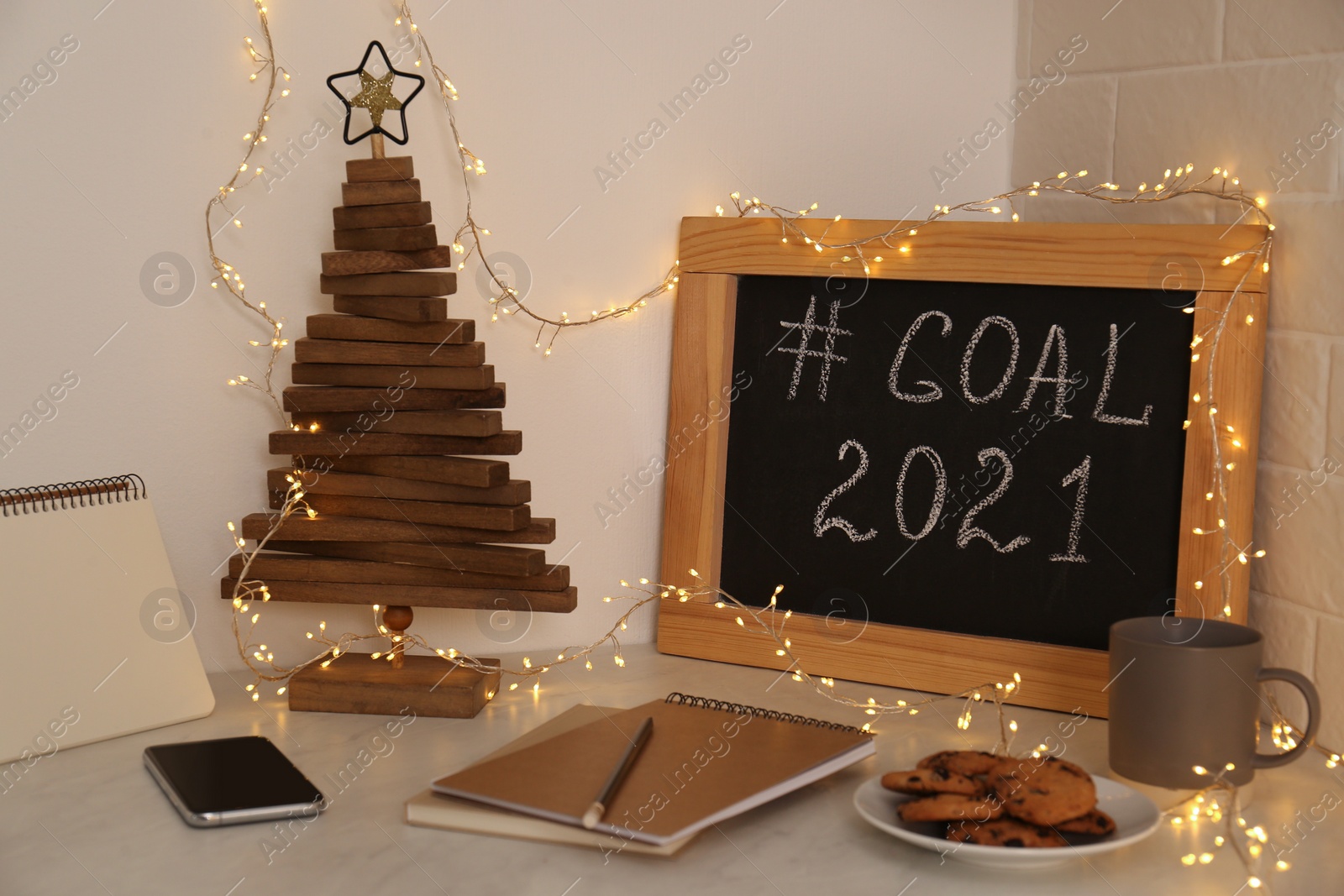 Photo of Chalkboard with hashtag Goal 2021, fairy lights near notebooks, smartphone and cookies on white table. New year targets