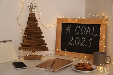 Photo of Chalkboard with hashtag Goal 2021, fairy lights near notebooks, smartphone and cookies on white table. New year targets