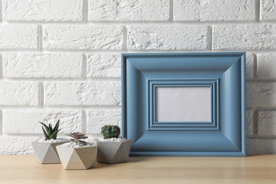 Photo of Blank frame and succulent plants on wooden table near white brick wall, space for design. Home decor