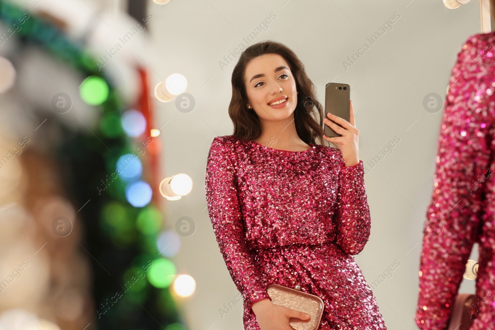 Photo of Beautiful young woman trying on stylish pink sequin dress and taking selfie near mirror in boutique. Party outfit