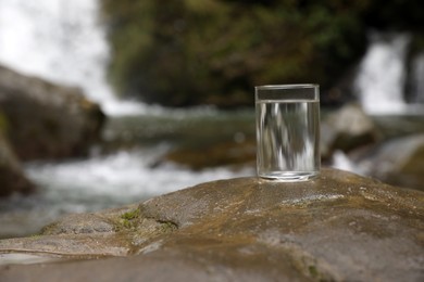 Glass of fresh water on stone near stream outdoors. Space for text