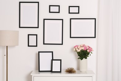 Photo of Empty frames hanging on white wall and chest of drawers with flowers indoors