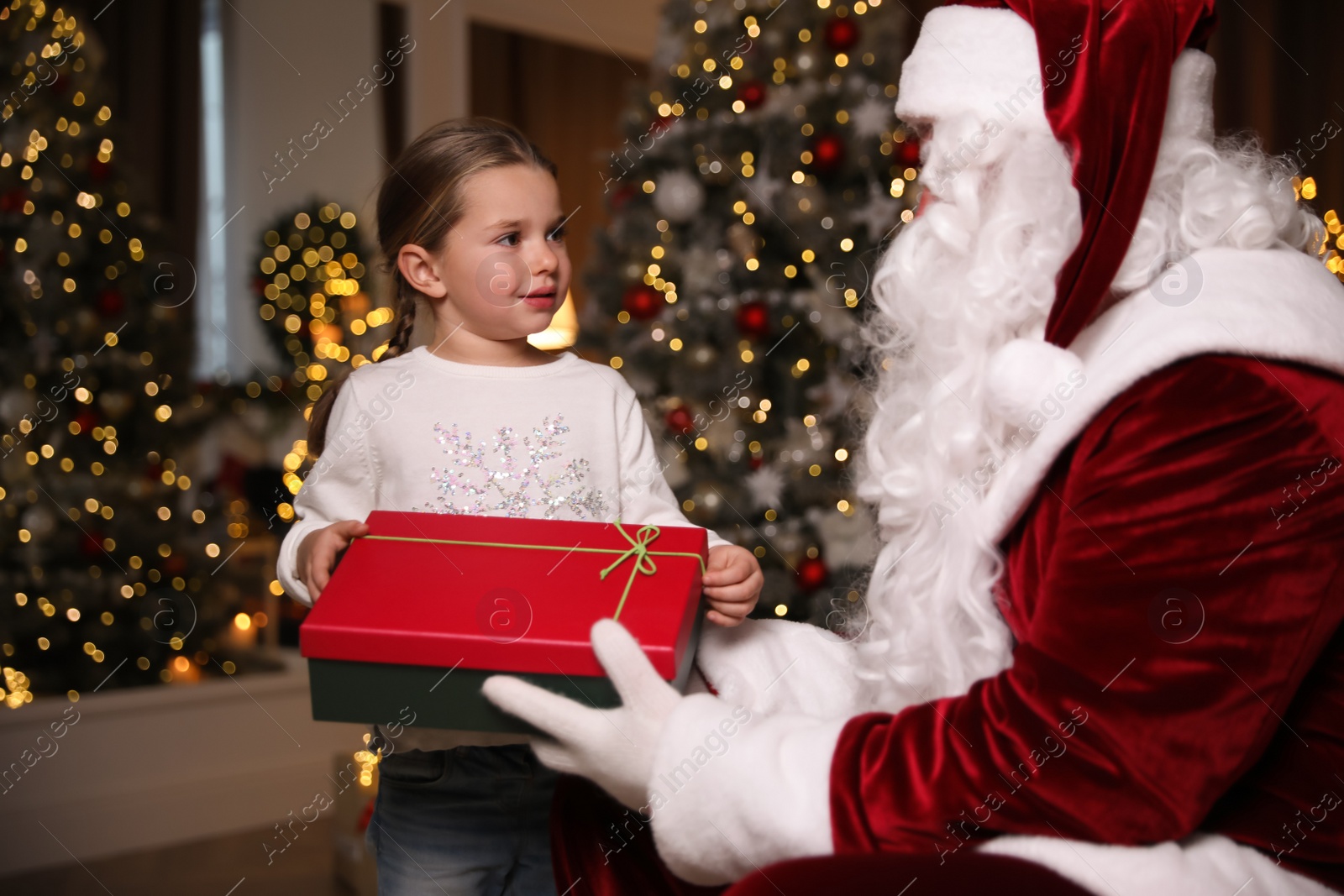 Photo of Santa Claus giving present to little girl in room decorated for Christmas