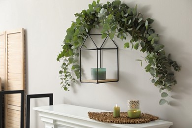 Photo of Beautiful garland made of eucalyptus branches and aromatic candle hanging above mantelpiece indoors