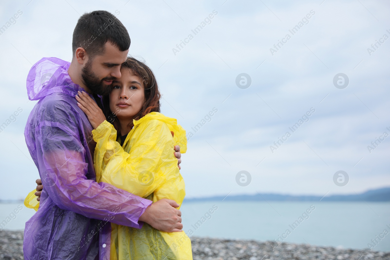 Photo of Young couple in raincoats enjoying time together under rain on beach, space for text