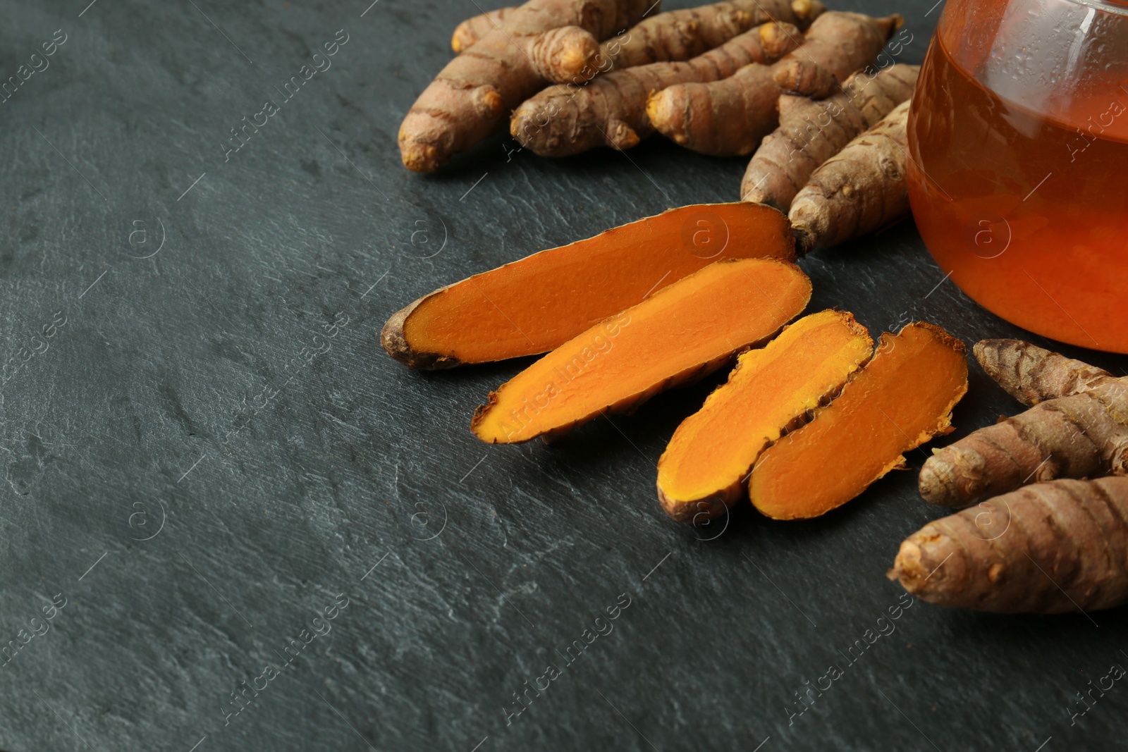 Photo of Glass cup of tasty tea, whole and cut turmeric roots on black textured table, space for text