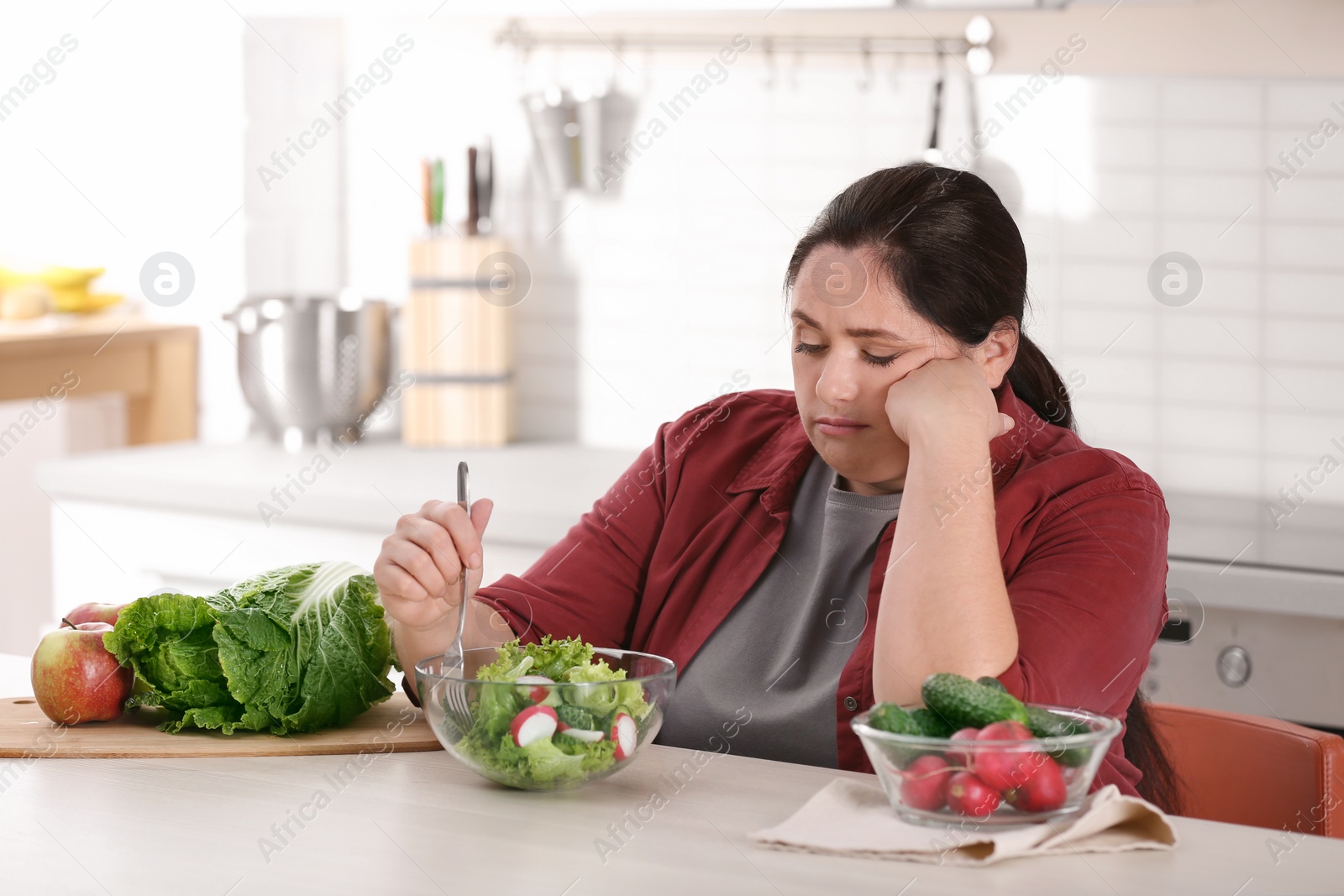 Photo of Unhappy woman eating vegetable salad at table in kitchen. Healthy diet