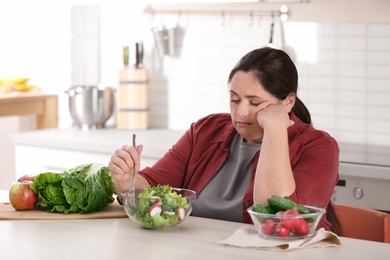 Photo of Unhappy woman eating vegetable salad at table in kitchen. Healthy diet