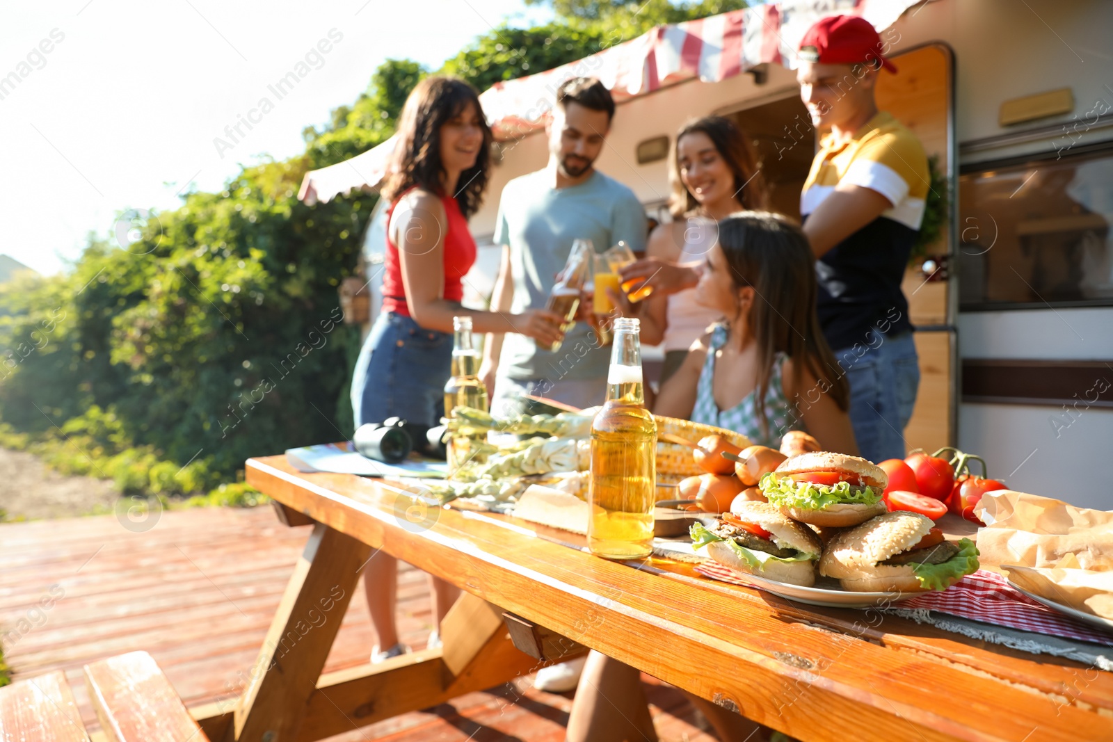 Photo of Friends toasting with bottles of drinks near motorhome, focus on table full of tasty food. Camping season