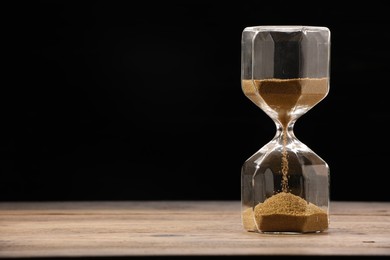 Photo of Hourglass with flowing sand on wooden table against black background. Space for text