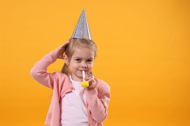 Photo of Birthday celebration. Cute little girl in party hat with blower on orange background, space for text