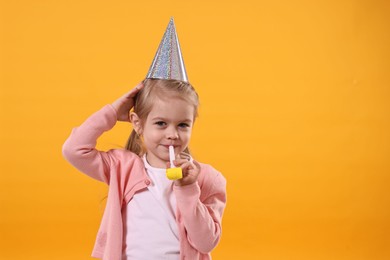 Birthday celebration. Cute little girl in party hat with blower on orange background, space for text