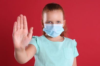 Photo of Little girl in protective mask showing stop gesture on red background. Prevent spreading of coronavirus