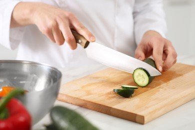 Photo of Professional chef cutting fresh cucumber at white table in kitchen, closeup