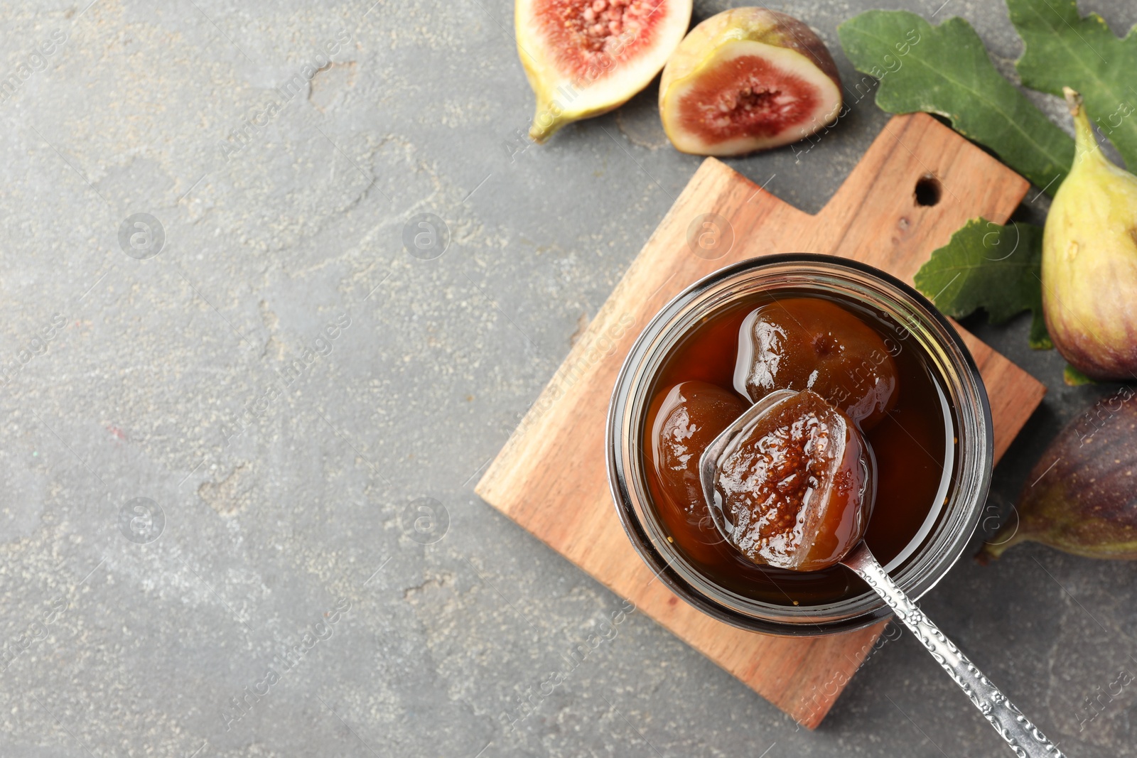 Photo of Jar of tasty sweet jam, fresh figs and green leaf on grey table, flat lay. Space for text