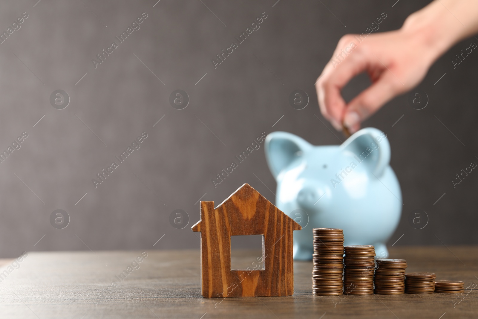 Photo of Woman putting coin into piggy bank at wooden table, focus on house model and money. Space for text