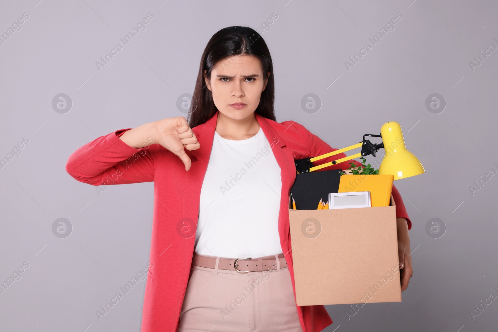 Photo of Upset unemployed woman with box of personal office belongings showing thumb down on grey background