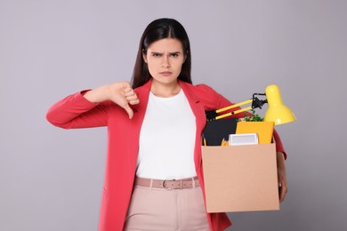 Photo of Upset unemployed woman with box of personal office belongings showing thumb down on grey background