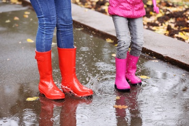 Mother and daughter wearing rubber boots splashing in puddle on rainy day, focus of legs. Autumn walk