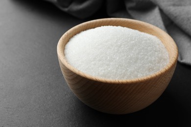 Photo of Granulated sugar in bowl on black table, closeup. Space for text