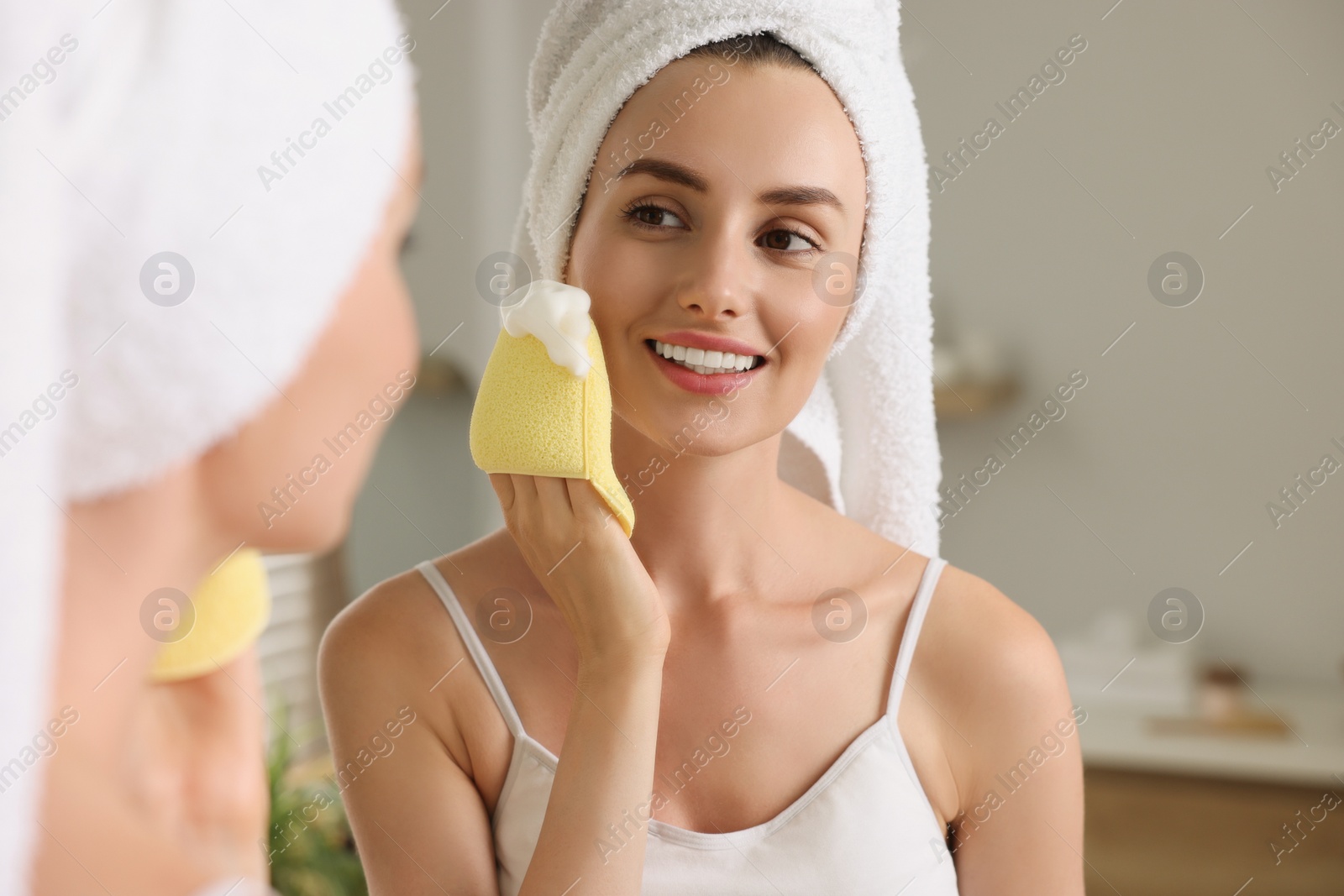 Photo of Happy young woman washing her face with sponge near mirror in bathroom