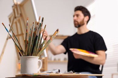Photo of Man painting in studio, focus on holder with brushes. Using easel to hold canvas