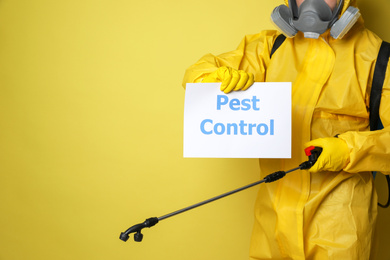 Man wearing protective suit with insecticide sprayer holding sign PEST CONTROL on yellow background, closeup. Space for text