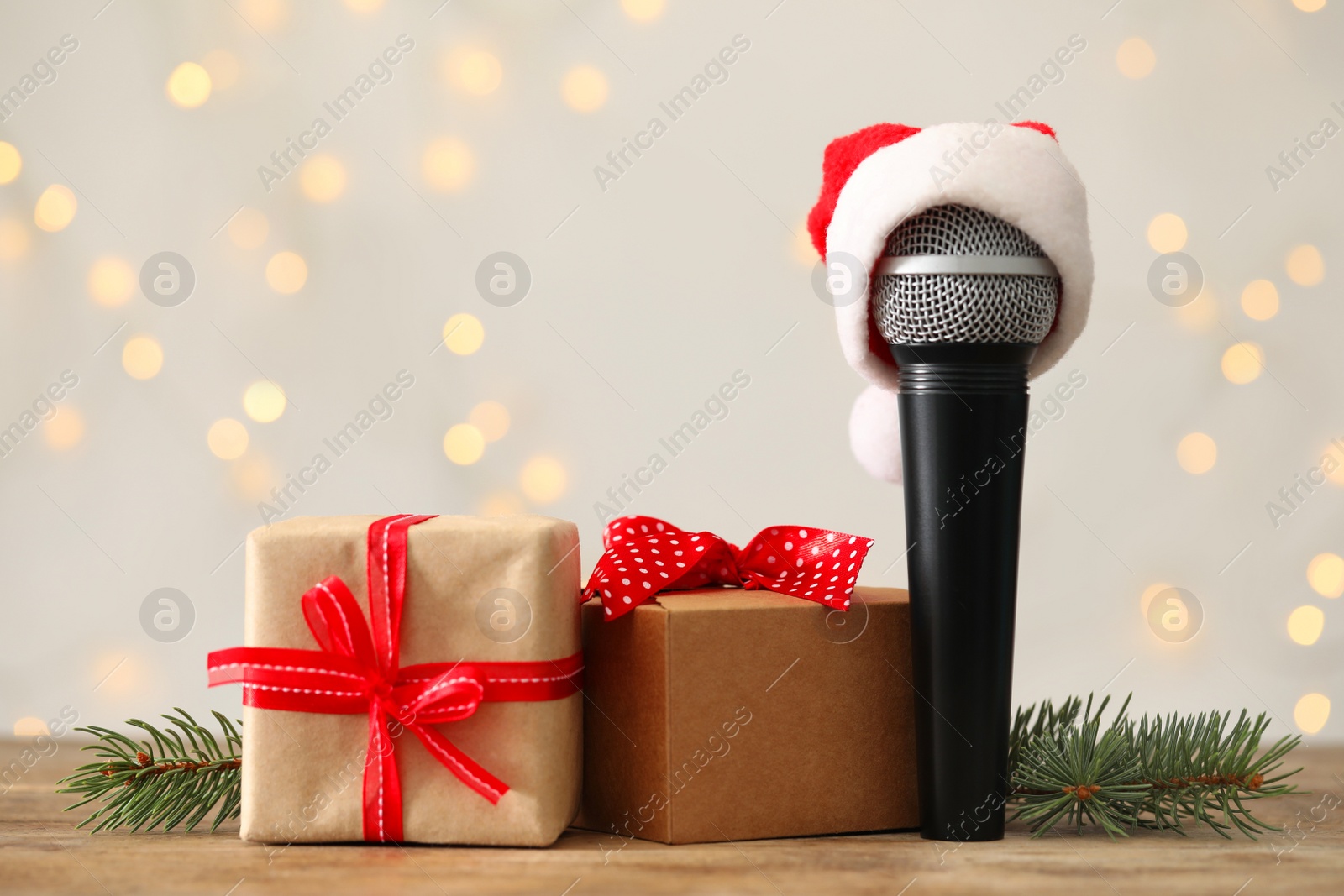 Photo of Microphone with Santa hat, gift boxes and fir branches on wooden table against blurred lights. Christmas music