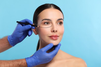 Image of Woman preparing for cosmetic surgery, light blue background. Doctor drawing markings on her face, closeup