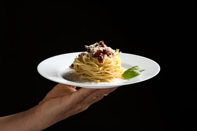 Woman holding plate of tasty spaghetti with sun-dried tomatoes and parmesan cheese on black background, closeup. Exquisite presentation of pasta dish