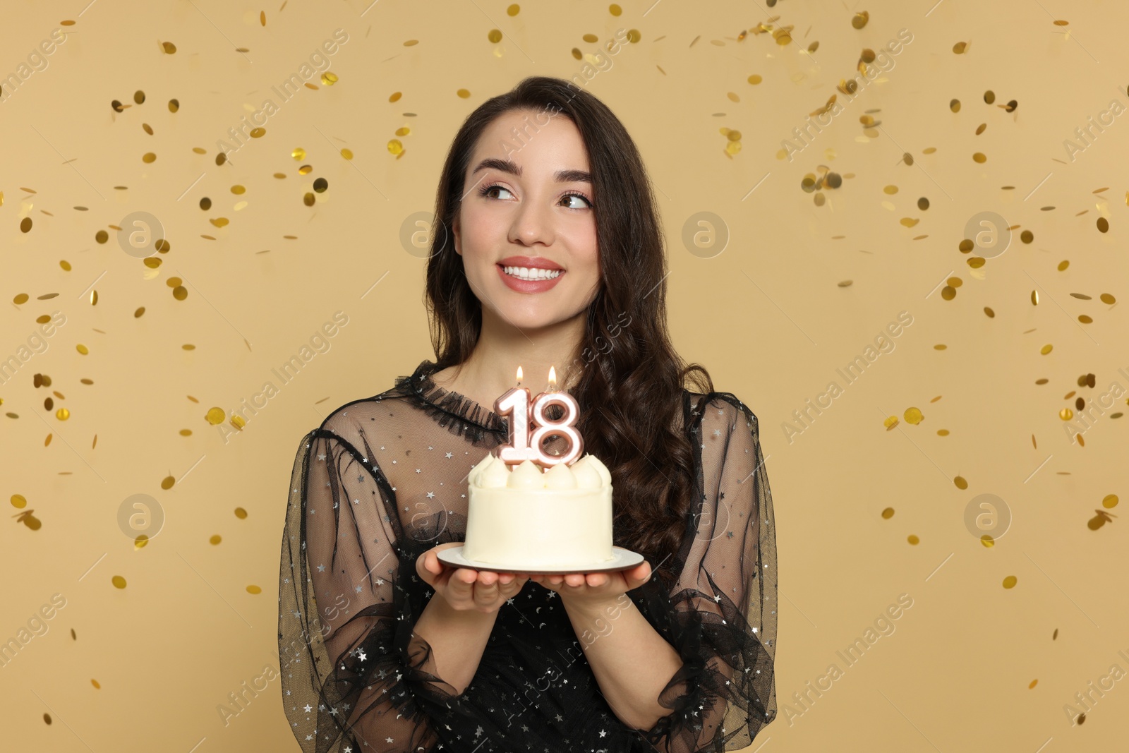 Photo of Coming of age party - 18th birthday. Woman holding delicious cake with number shaped candles and looking at falling confetti against beige background