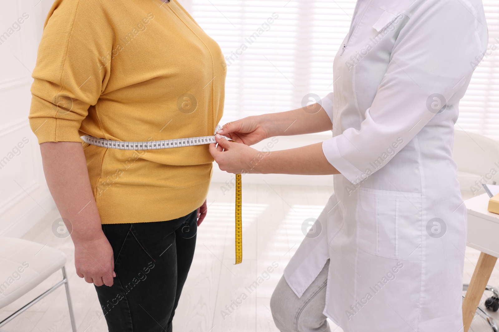 Photo of Nutritionist measuring overweight woman's waist with tape in clinic, closeup