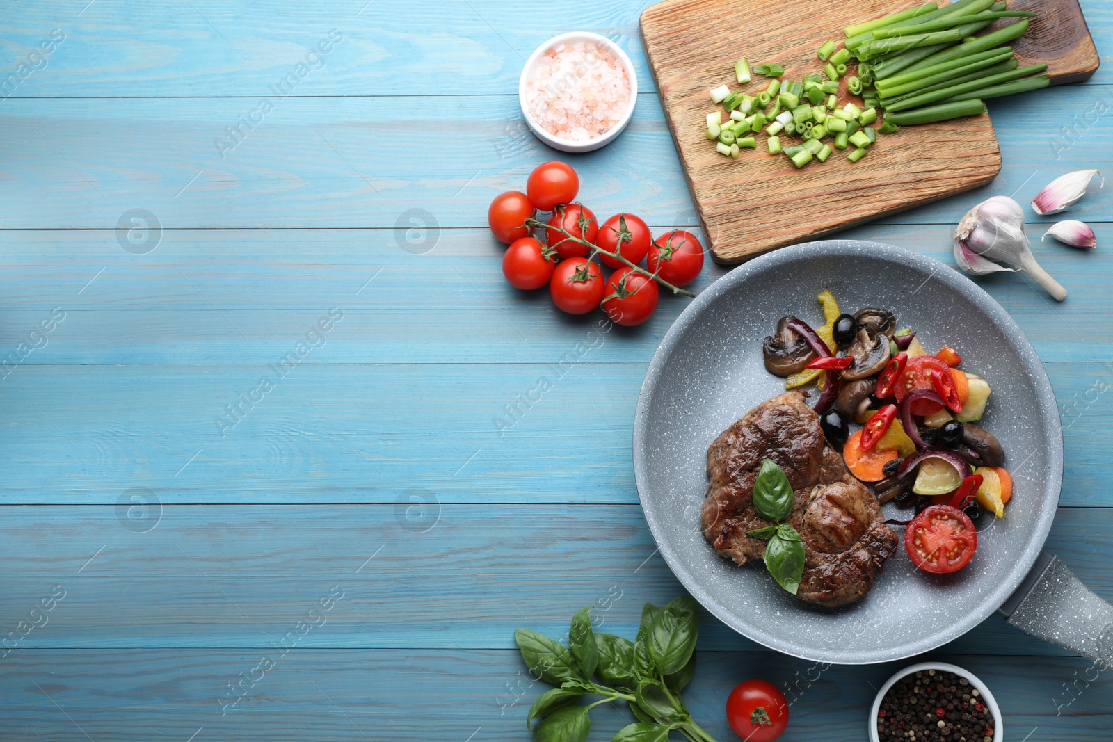 Photo of Tasty fried steak with vegetables in pan and ingredients on light blue wooden table, flat lay. Space for text