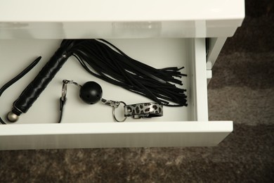 Black whip and ball gag in drawer indoors, above view. Sex toys