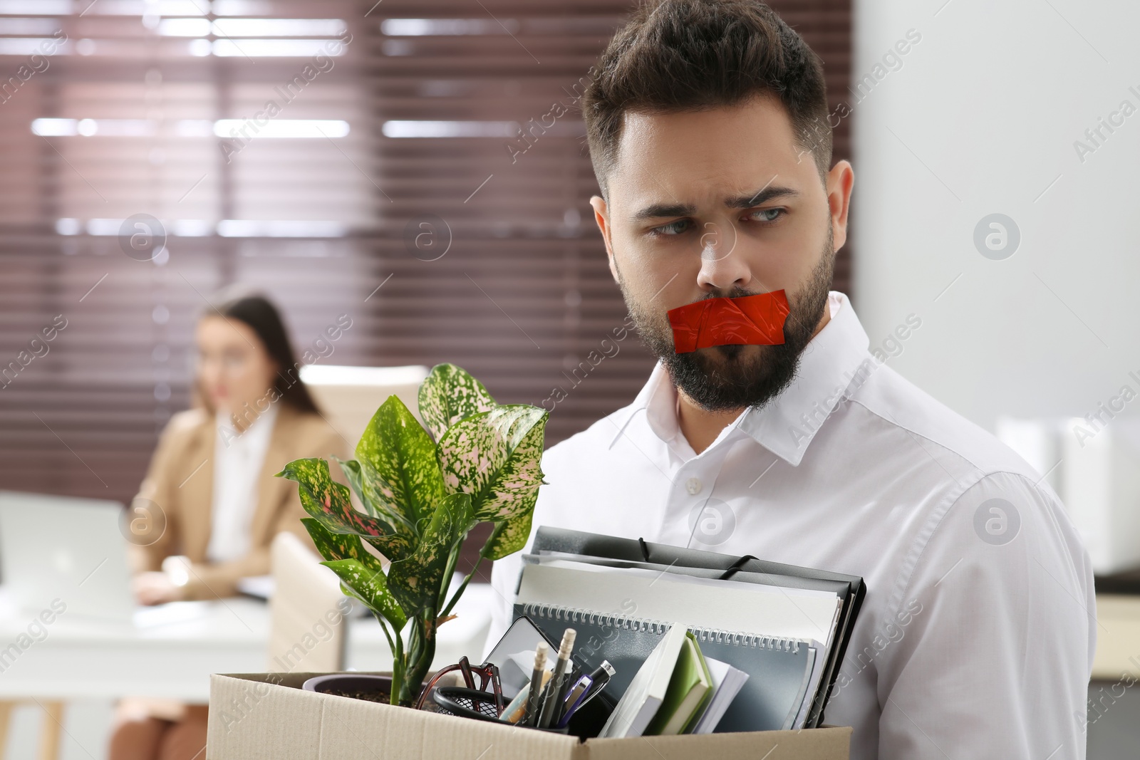 Image of Upset dismissed man with taped mouth carrying box with personal stuff in office. Speech censorship