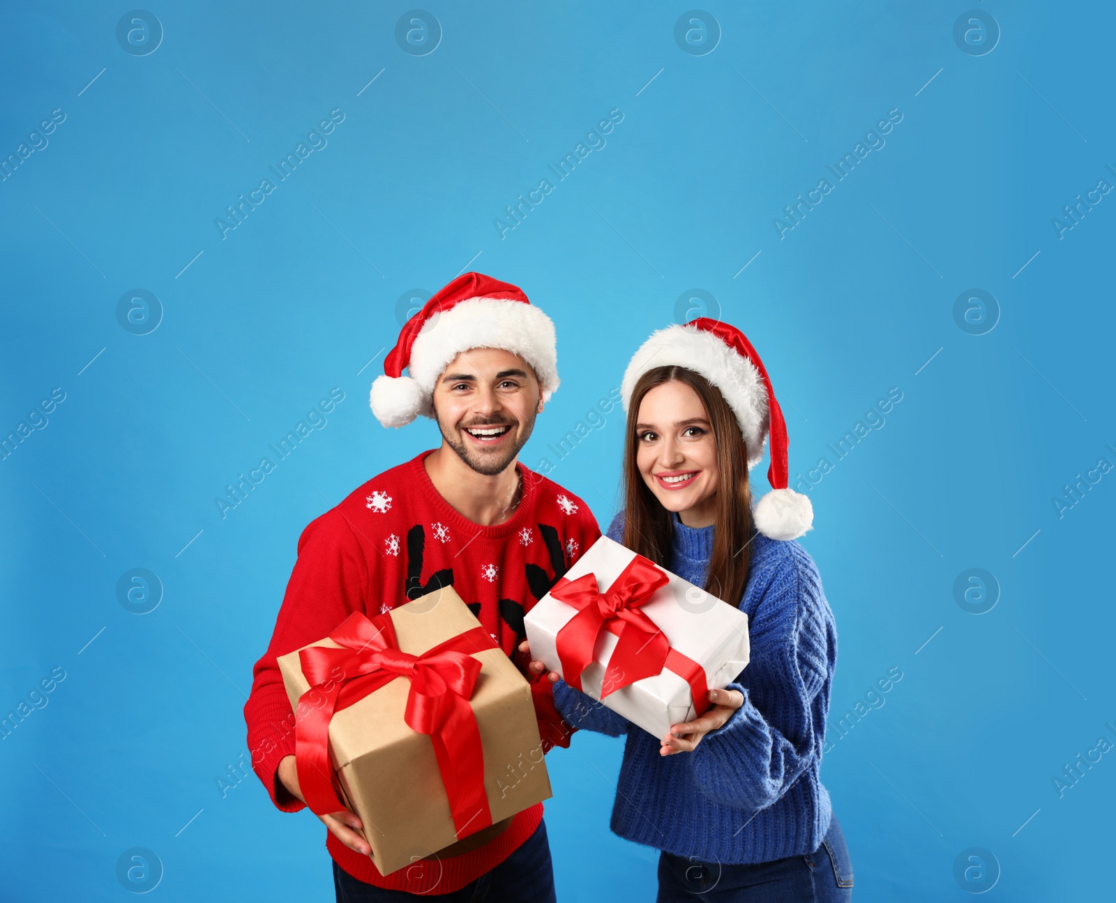 Photo of Couple in Christmas sweaters with Santa hats and gift boxes on blue background