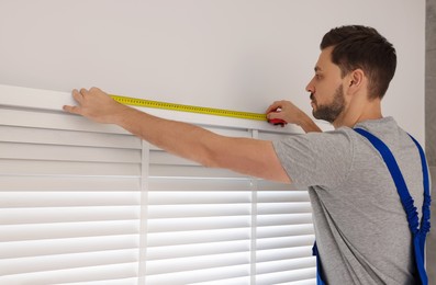 Worker in uniform using measuring tape while installing horizontal window blinds indoors