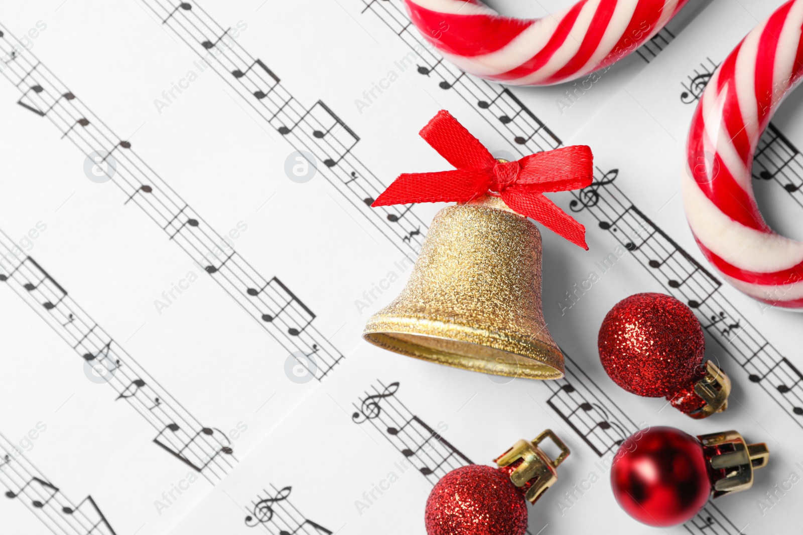 Photo of Golden shiny bell with red bow, candy canes and Christmas baubles on music sheets, flat lay and space for text
