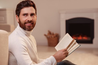 Handsome man reading book near fireplace in room