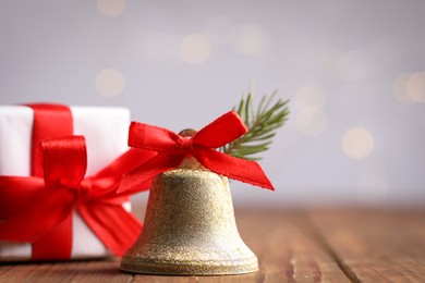 Photo of Bell with red bow and gift box on wooden table against blurred background, closeup. Christmas decor