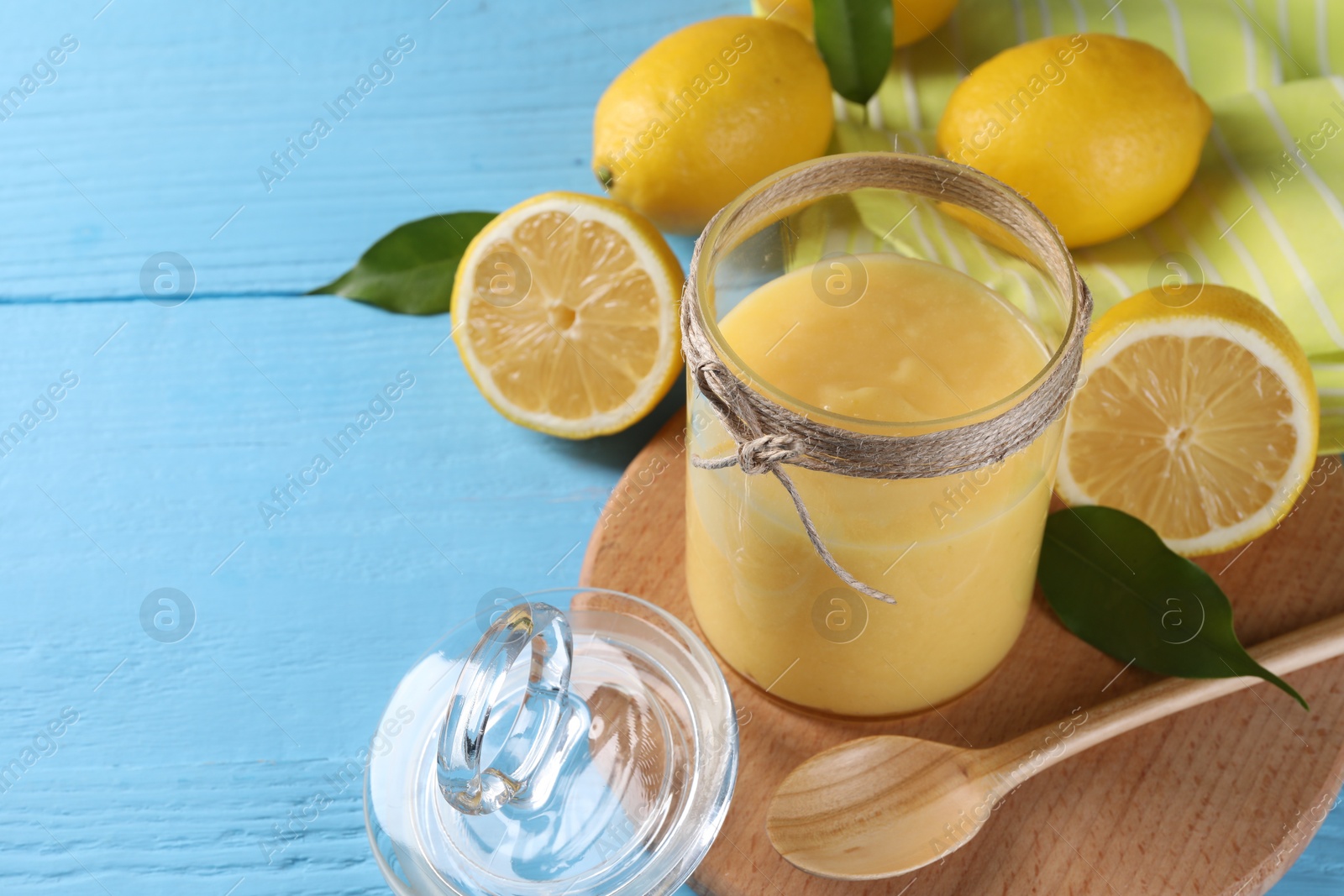 Photo of Delicious lemon curd in glass jar, fresh citrus fruits, spoon and green leaves on light blue wooden table, space for text