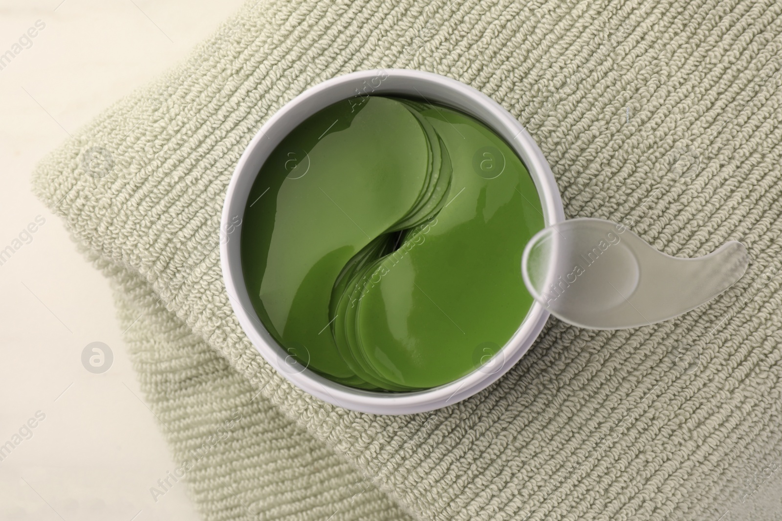 Photo of Jar of under eye patches with spoon and towel on white background, top view. Care products