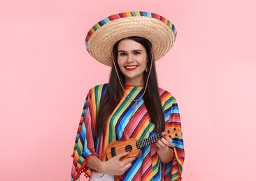 Young woman in Mexican sombrero hat and poncho playing ukulele on pink background