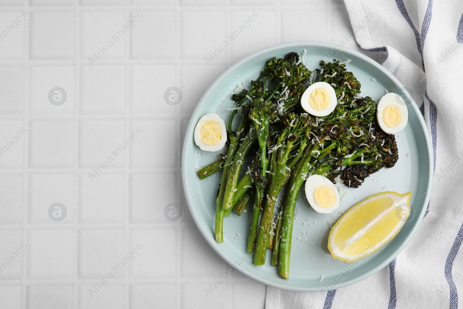 Photo of Tasty cooked broccolini with cheese, quail eggs and lemon on white tiled table, top view. Space for text